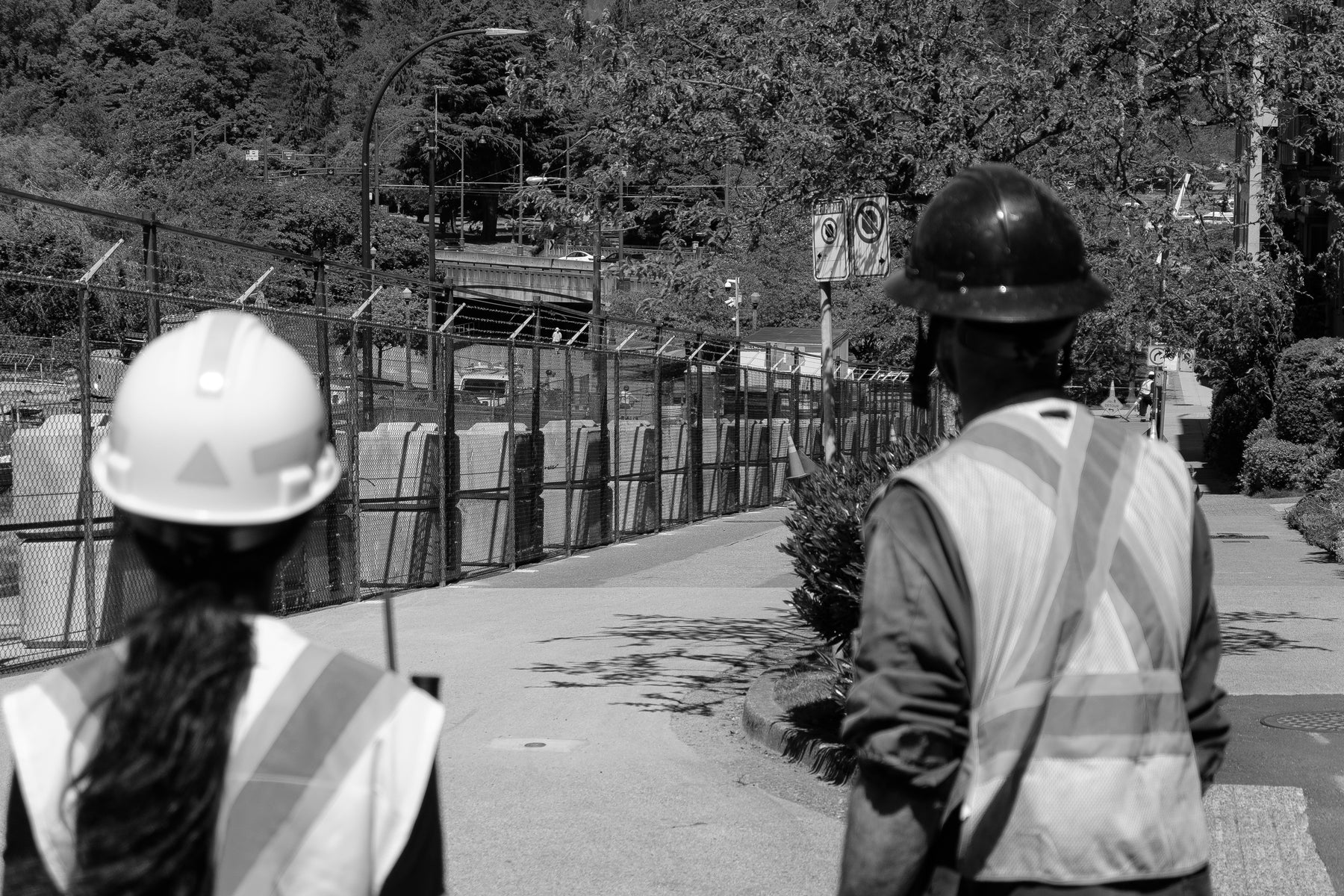 Two construction workers wearing hard hats and safety vests walk along a fenced path near a roadway
